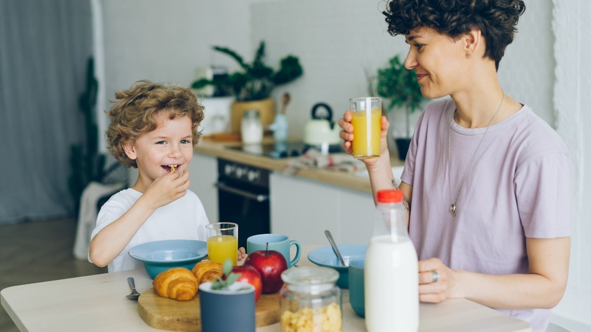 Young woman drinking orange juice while son eating cereal at table in kitchen