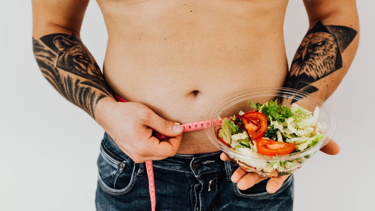 Tattooed man measuring waist while holding fresh salad, symbolizing healthy lifestyle and weight management.
